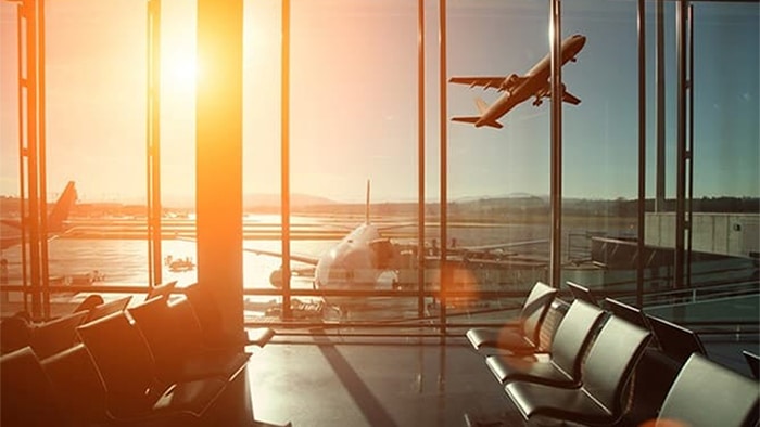 A photo taken inside an airport terminal. It shows empty seats and the window beyond shows airplanes on the tarmac. One plane is in midflight, taking off from the runway.