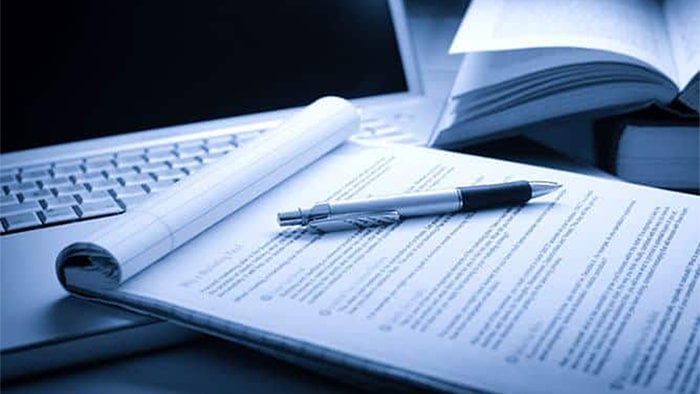 A pen is on top of documents propped in front of an open laptop.