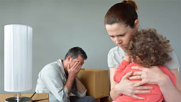 A woman is hugging a child, with a man covering his face with his hands in the background. He sits next to a packed box.