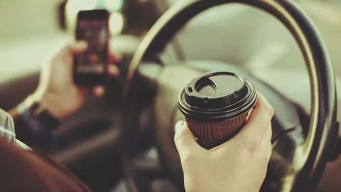An unidentified person sitting behind the wheel of a car with a cellphone in one hand and a coffee in the other.