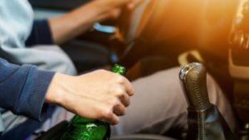 The focus is on a driver in the driver's seat, holding a beer with one hand and the steering wheel in the other.