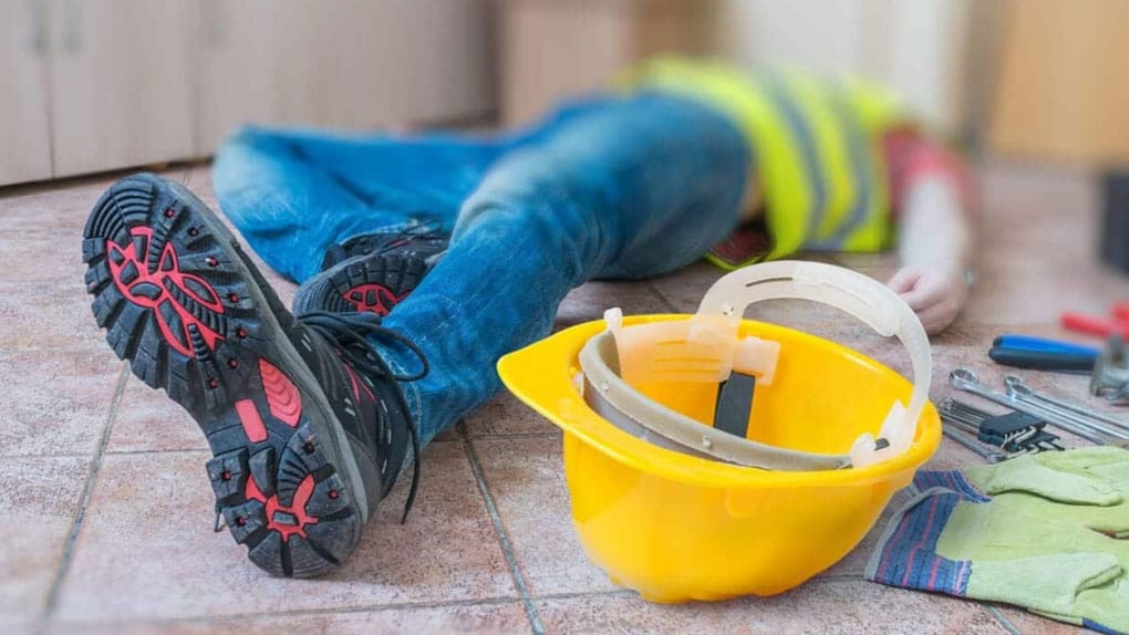 The focus is on the lower half of an injured worker lying on the ground; his yellow hard hat is next to him.