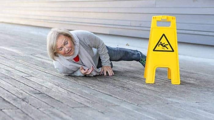 An elderly woman has fallen; she lies next two a yellow caution sign.