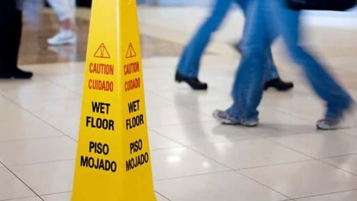 A yellow caution: wet floor sign. In the background are blurred images of people walking by the sign.