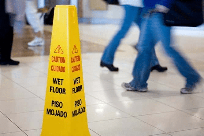 A yellow caution: wet floor sign. In the background are blurred images of people walking by the sign.