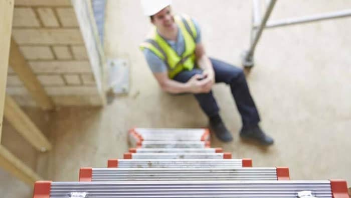Focus is on the top of a ladder. Below, on the ground, out of focus is a worker in a yellow vest and hard hat, clutching his knee.