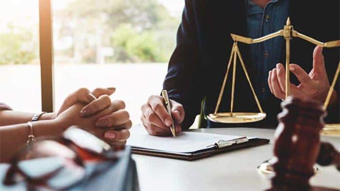 Focus is on a lawyer discussing the case with their client. On their desk are paperwork, the scales of justice, and a gavel.