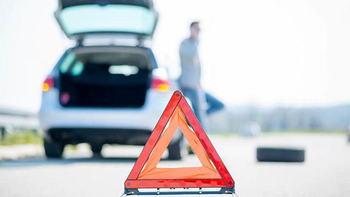 Focus is on a road sign, alerting danger. In the background is a broken-down vehicle with a man on a cell phone next to it. Both are out of focus. A tire lays in the road.