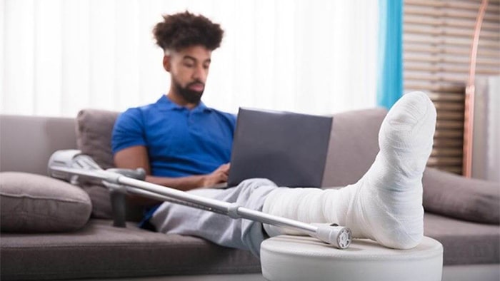 A man with a broken leg rests his casts on a table as he sits on the couch with an open laptop in his lap. His crutch is propped up beside him.