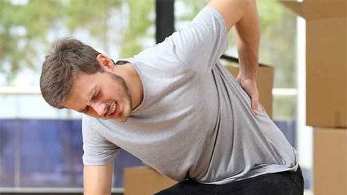 An injured man is bending over, holding his back as he leans against a box he is trying to lift. More boxes are stacked in the background.