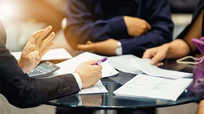 The focus is on the paperwork and hands of three people, meeting around a circular table and discussing.