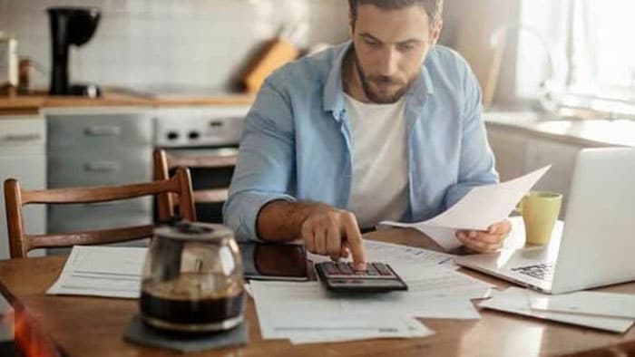 A man sits at his table with a calculator, laptop, and bills surrounding him.