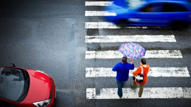 An aerial view of two pedestrians crossing a crosswalk with an umbrella as cars drive by.