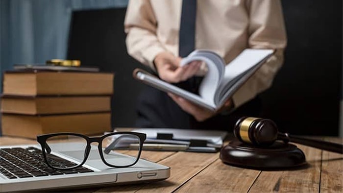 The focus is on a laptop with a pair of glasses resting on top, a gavel, and paperwork. A stack of books is in the background, along with a blurred out view of a lawyer reading a book.
