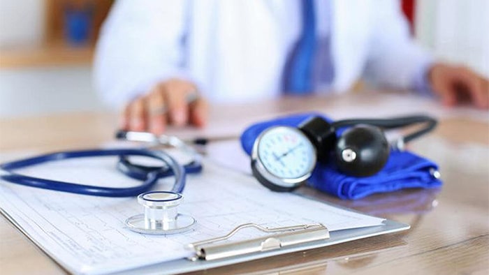 A stethoscope and a blood pressure cuff sit on top of the paperwork. A doctor is out of focus in the background.