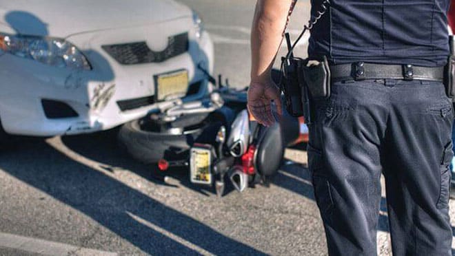 The rear view of a police officer standing near the scene of a motorcycle accident. The motorcycle is partially trapped beneath the car.