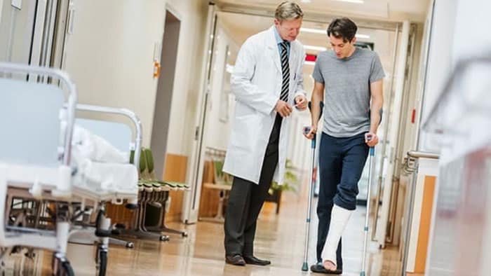 A doctor is working with an injured patient who has a cast on their leg as they try to walk down a hospital hallway with crutches.