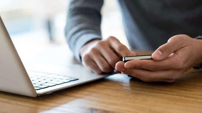 A man is typing on his cell phone next to an open laptop.