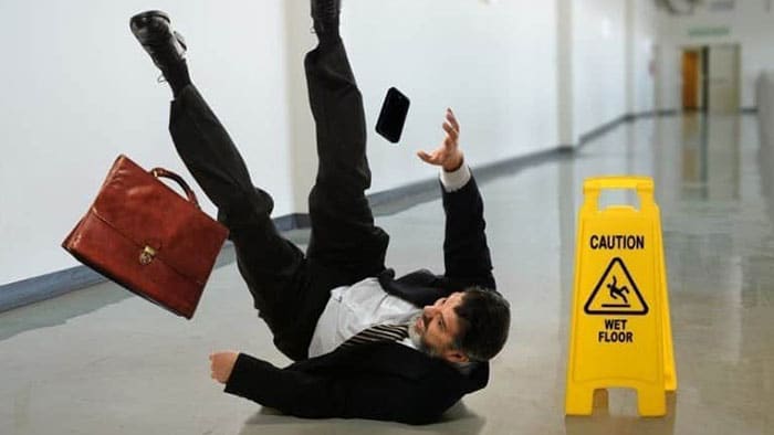 A man is upside down with his feet in the air, mid-fall, after slipping next to a caution: wet floor sign.