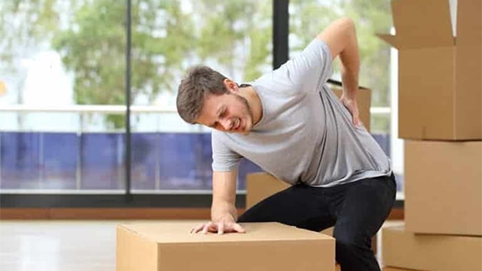 An injured man is bending over, holding his back as he leans against a box he is trying to lift. More boxes are stacked in the background.