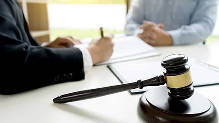 The focus is on a gavel. In the background, an attorney is filling out paperwork for their client.