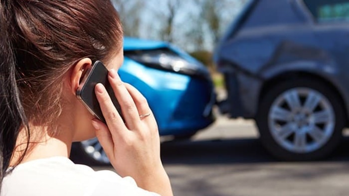 The focus is on a woman calling the police after an accident. The damaged vehicles are in the background.