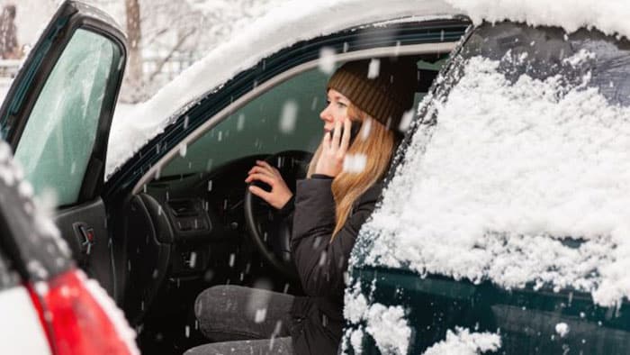 A woman is in the driver's seat with her door open while on her cell phone. Her car is covered in snow.