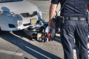 The rear view of a police officer standing near the scene of a motorcycle accident. The motorcycle is partially trapped beneath the car.