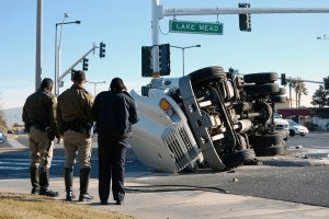 Three people stand at a corner; in the roadway is a tipped-over tractor-trailer.