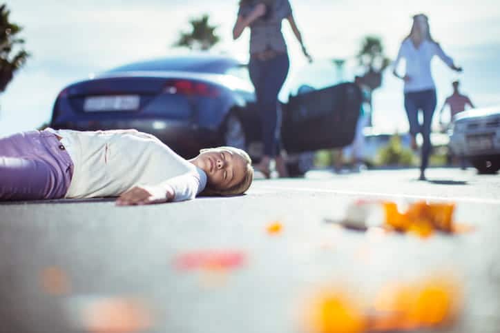 An unconscious woman lying in a parking lot after being hit by a car. People are running toward her in the background, along with the car that has stopped.