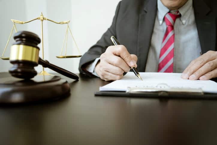 Focus is on a lawyer working on paperwork at his desk. There is a gavel and a scales of justice next to him.