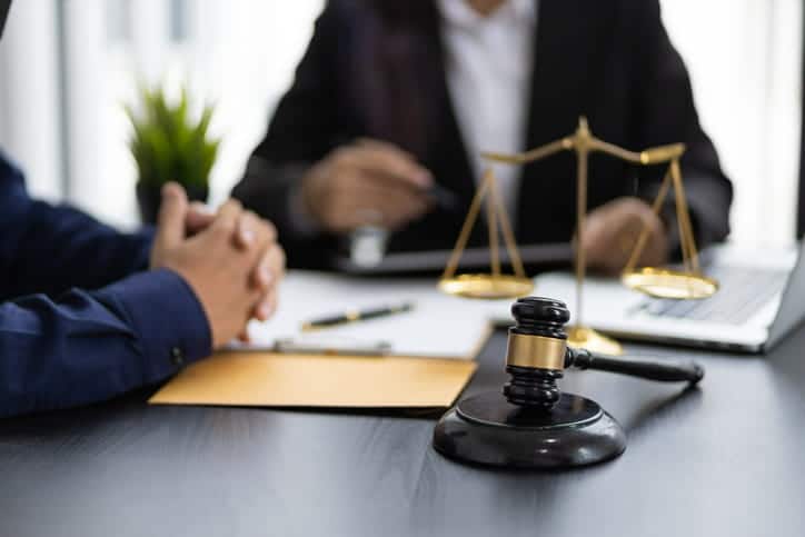 Focus is on a gavel sitting on a desk, with the scales of justice behind it. Behind the objects are a lawyer and a client, out of focus, as they discuss the case.