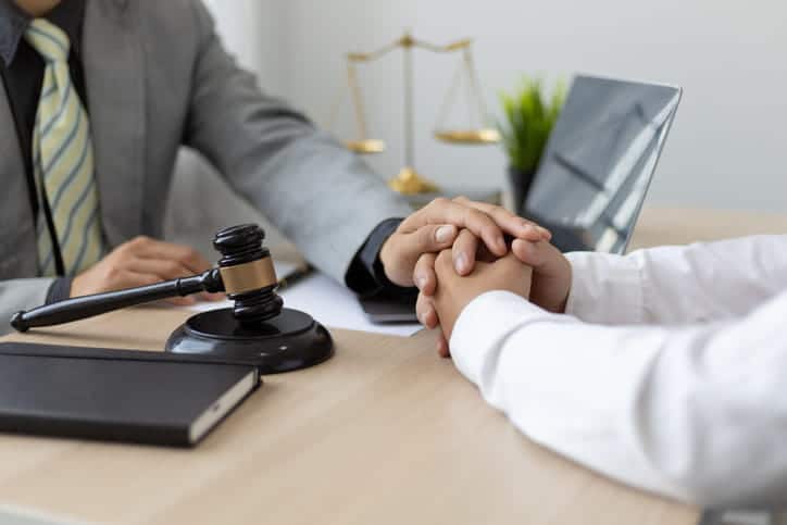 Focus is on a lawyer consoling their client, with their hand resting over the hurt individual. On their desk is an open laptop, paperwork, a gavel, and the scales of justice.