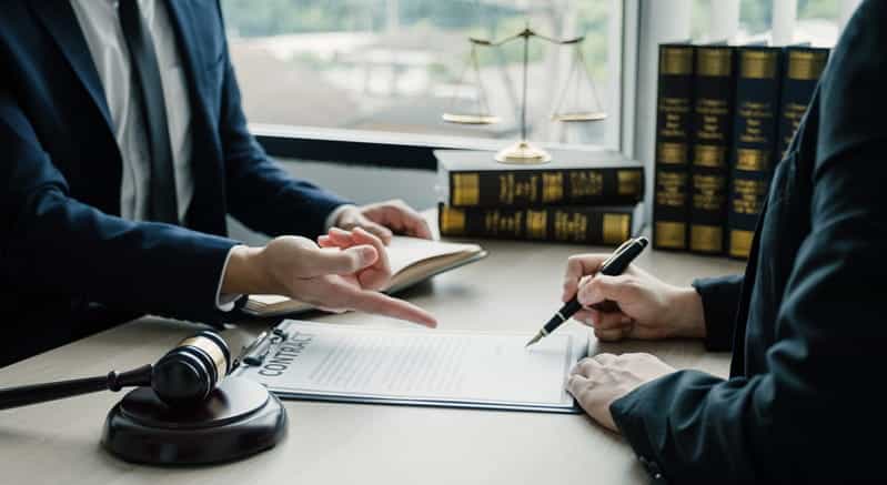 Focus is on a lawyer showing their client where to sign on a document. Next to them is a gavel and a pile of books.