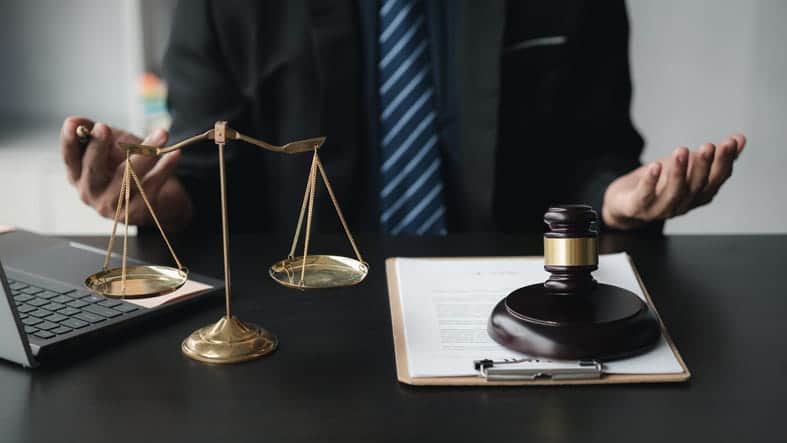 Focus is on a lawyer sitting at his desk, can only see his hands. On his desk is an open laptop, paperwork, a gavel, and a scales of justice.