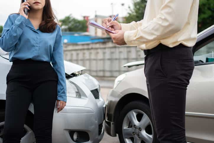 Focus is on two crashed vehicles. There's a woman on her cellphone standing in front of one, and then a man with a clipboard taking her statement.