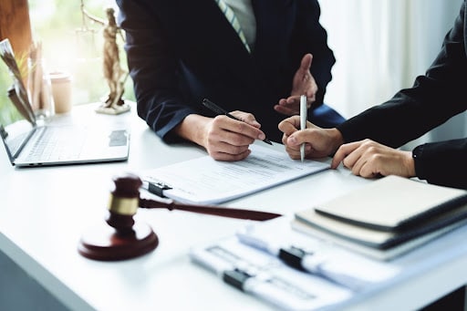 two lawyers are discussing their clients paperwork. a gavel and paperwork are on their desk, along with an open laptop.