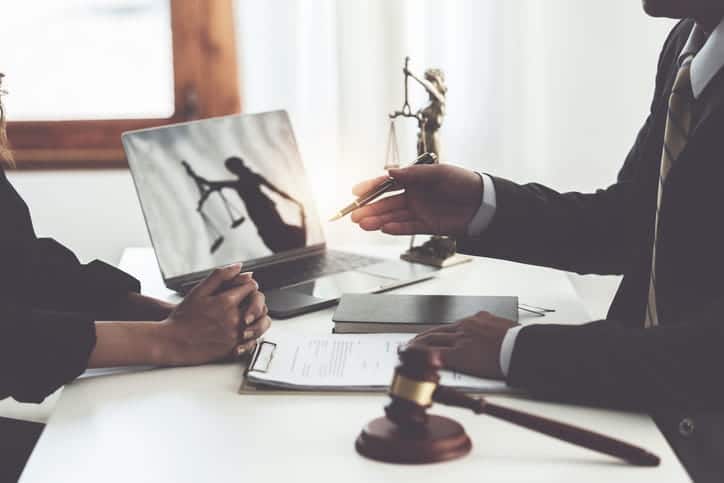 A Vegas truck accident lawyer speaks with their client about their case. The focus is them at the lawyer's desk, on it is an open laptop, a gavel, a Lady Justice statue, and paperwork.