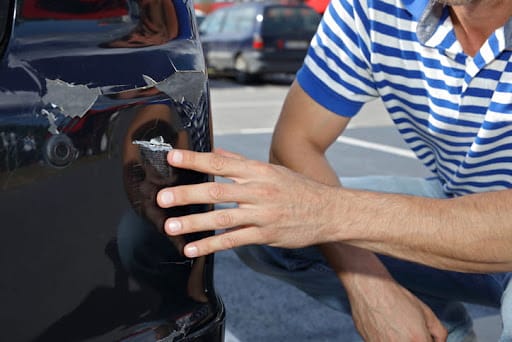 The focus is on a man kneeling, inspecting the damage on his vehicle after a parking lot accident.