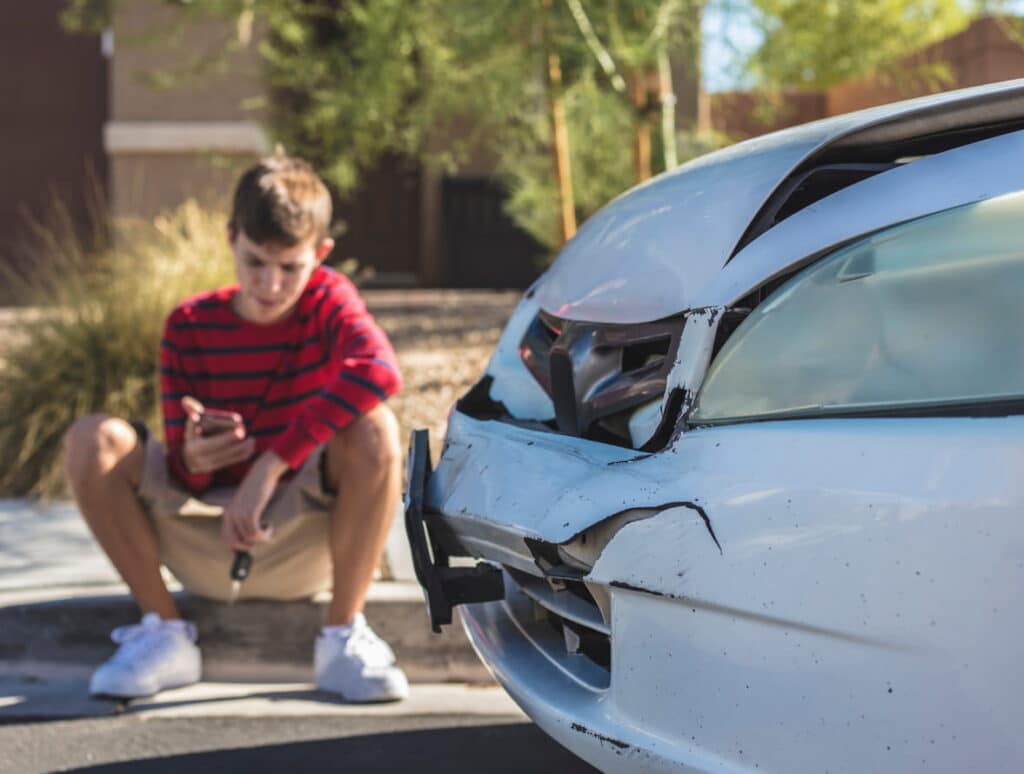 A young man sits on the curb as he waits for help after a minor car accident. In the photo, the front end of the vehicle shows damage.