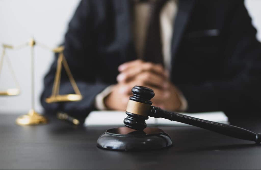 Focus is on a gavel that sits on a car accident lawyer's desk. In the background, the lawyer sits with his hands clasped together and the scales of justice is next to him.