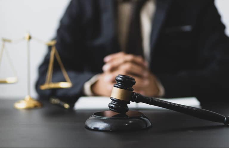 Car Accident Claim: Speeding 2 Focus is on a gavel that sits on a car accident lawyer's desk. In the background, the lawyer sits with his hands clasped together and the scales of justice is next to him.
