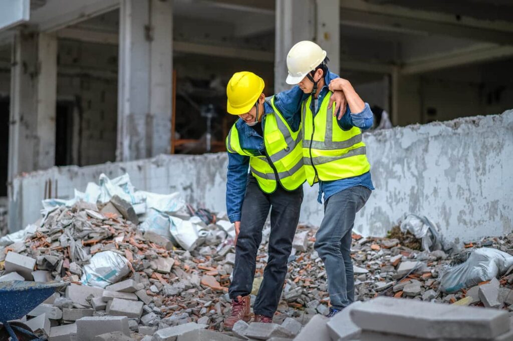 Two-construction-workers-in-yellow-vests-one-is-holding-the-other-up-as-they-walk-through-rubble.-The-one-worker-has-suffered-a-workers-comp-injury