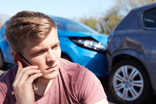 A young man on his cellphone after being in a minor car accident. The damaged vehicles are in the background after being in a rear-end collision.