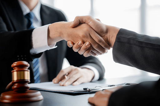 A personal injury lawyer shaking their client's hand after an injury lawsuit settlement has occurred. On the desk is a gavel and paperwork.