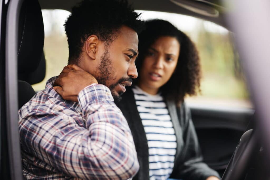 A man holds his neck after suffering whiplash in a car accident. His wife is in the driver's seat looking at him in concern.