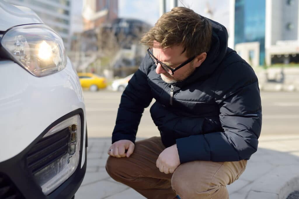 A man is kneeling by the front of a vehicle, inspecting for rental car damage.