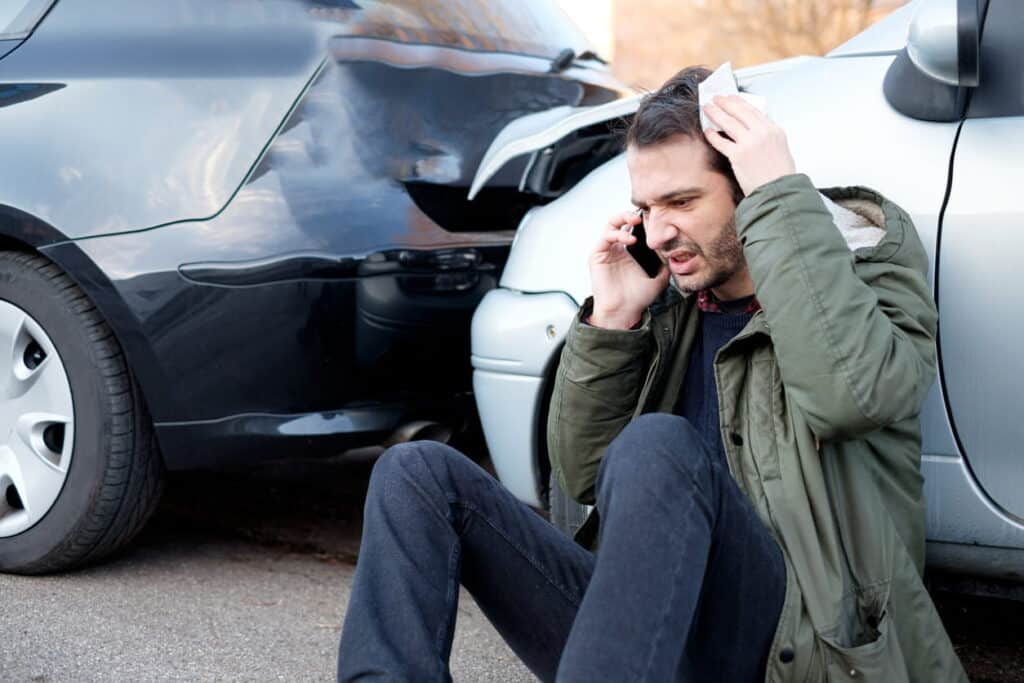 A man upset on the phone as he sits on the ground against his vehicle that has recently been in a read-end collision. It's still touching the other vehicle in the background.