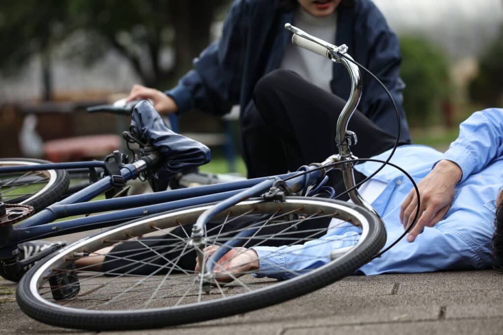 An injured person lying on the ground after being in a bicycle accident. His bicycle is next to him, as a woman checks on him.