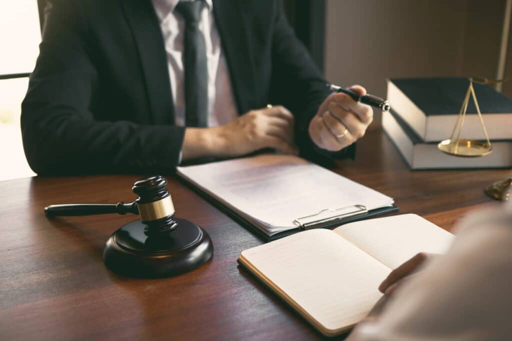 Focus is on a car accident lawyer working at his desk with a client. On his desk is paperwork and a gavel.